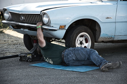 A mechanic working under a classic car on the pavement during the day.